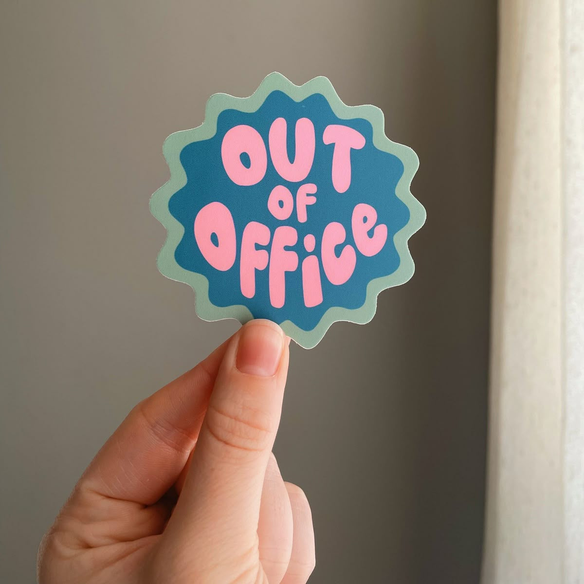 Hand holding a 'Out of Office' sticker with a blue and pink design.