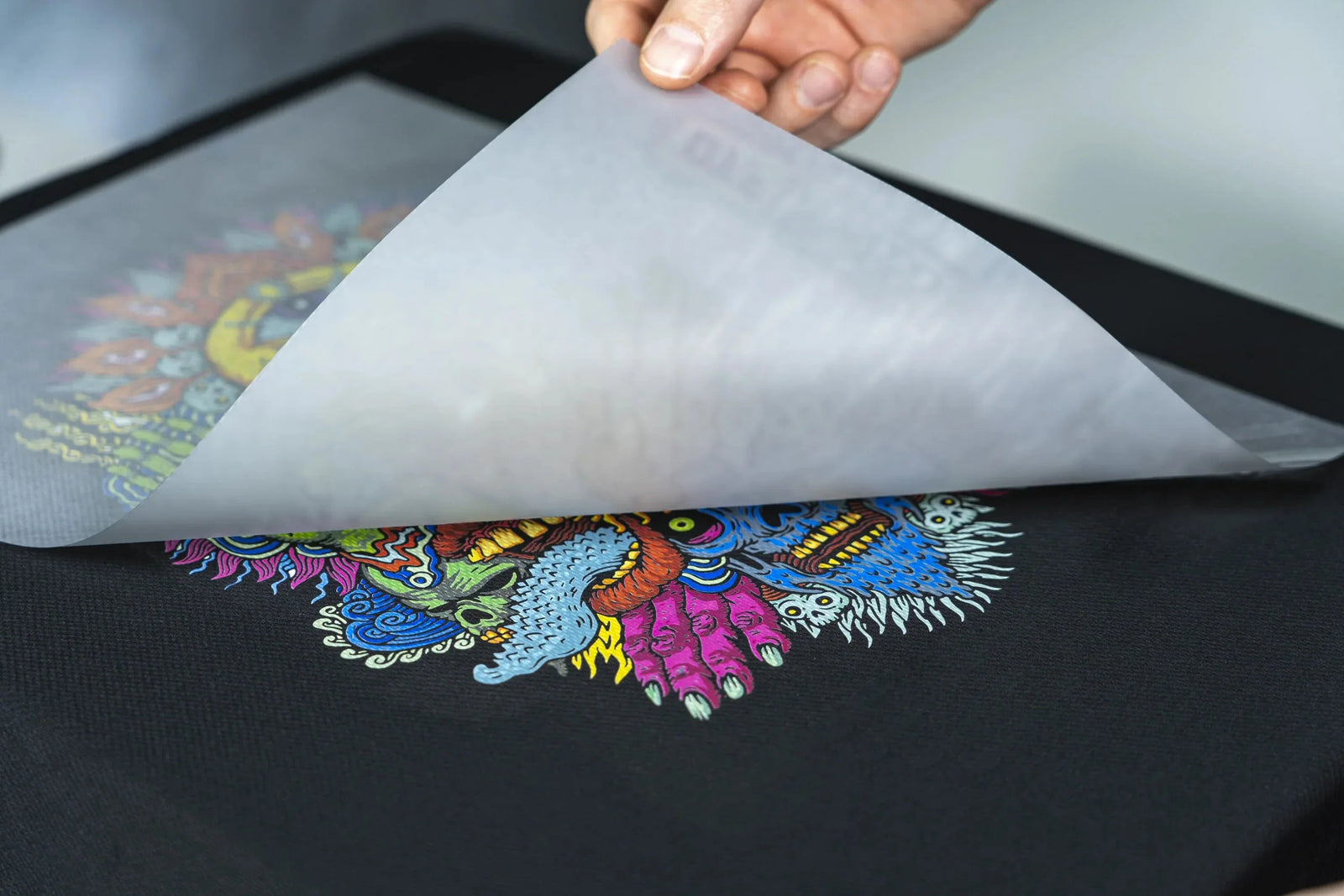 Hand peeling away transfer paper from colorful monster face design on black fabric