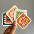 A hand holds four square cards with rounded corners, each featuring a colorful geometric medallion pattern, against a plain gray background.