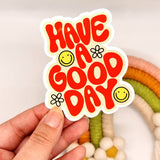 Sticker with 'Have a Good Day' text held by a hand against a neutral background